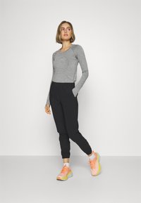 Tricou lung gri cu mâneci, pantaloni jogger negri cu buzunare și pantofi sport roz cu talpă galbenă, stând pe un fundal neutru.
