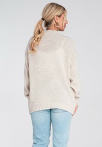 Jasno beżowy sweter z dzianiny o luźnym kroju, opadłych ramionach, teksturowanej włóczce i prążkowanym wykończeniu. Połączony z jasnoniebieskimi spodniami w taperze.