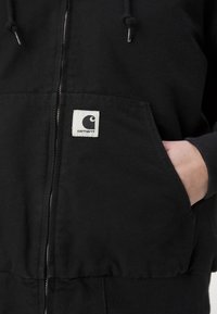 Black zip-up jacket made of cotton blend fabric. Features a large front pocket and a square logo patch with a curved design on the chest.
