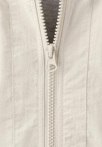 Svetlo bež jakna z zadrgo, ki ima gladko teksturo tkanine, sredinsko zadrgo z vrvico iz plastike in navpične šive.