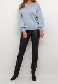 Femme portant un pull léger à manches longues bleu clair, un pantalon en cuir noir et des bottes noires à talons montant jusqu'aux genoux, posant devant un fond uni.