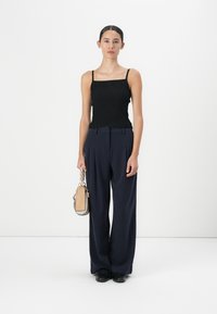 Black ribbed tank top, high-waisted navy pleated trousers, and a small beige handbag with a chain strap. Textured and tailored look.