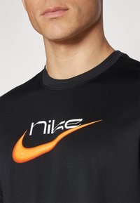 Detailní záběr na osobu v černém tričku s bílým logem "nike" a oranžovým logem Nike swoosh na hrudi.