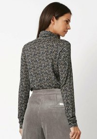 Langärmliges, figurbetontes Rollkragenoberteil mit einem schwarz-weißen, wirbelnden Muster. Weicher, elastischer Stoff mit hohem Kragen. Kombiniert mit grauer Cordhose.