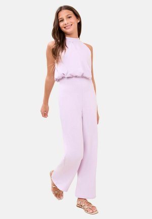 Niña sonriente, vestida con un mono sin mangas de color púrpura claro y sandalias planas beige con tiras, de pie frente a un fondo liso.