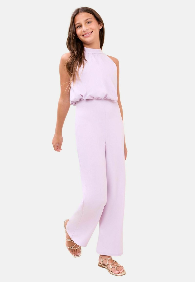 Fiatal lány mosolyog, ujjatlan halványlila overált és bézs színű, pántos lapos szandált visel, sima háttér előtt áll.