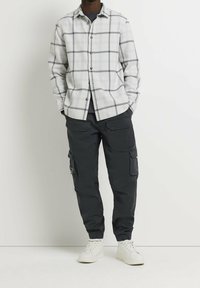 Camicia a quadri grigia e bianca, abbinata a pantaloni cargo neri con molte tasche. Il modello indossa sneakers bianche.