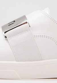 Vita lädersneaker med ett brett elastiskt band och en metallaccent. Slät textur och minimalistisk design med en platt sula.