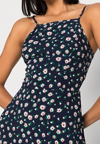 Žena v šatech bez rukávů tmavě modré barvy s tenkými ramínky a bílým a červeným květinovým vzorem s kopretinami.