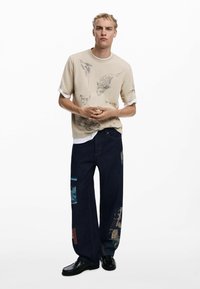 Beige short-sleeve shirt with graphic print; dark denim jeans with patchwork details; black shoes.