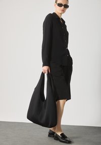 Svart oversize hobo-väska med slät textur, breda remmar och rundad botten. Bärs tillsammans med en svart knapp-up skjorta och shorts.