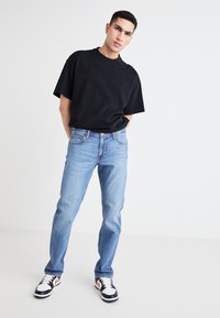 Črna bombažna majica, svetlo modri denim kavbojke in črno/rdeči športni čevlji. Kavbojke ravnega kroja z blagim obrabljenim videzom, majica s kratkimi rokavi.