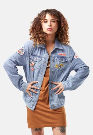 Femme aux cheveux bouclés portant une veste en jean bleu clair avec des patchs sur une robe marron, debout les mains sur les hanches devant un fond blanc.