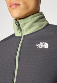 Sivi zip-up jakna s zelenim polarom oko vrata, s uzorkom u obliku mreže i bijelim logotipom na prsima. Detalj zatvarača je vidljiv.