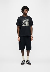 Μαύρο βαμβακερό T-shirt με γραφικό σχέδιο, συνδυασμένο με μαύρο σορτς. Το μοντέλο φοράει μαύρα αθλητικά παπούτσια και κρεμ κάλτσες. Απλό φόντο.