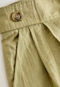 Gros plan sur un pantalon en tissu beige avec un devant plissé et un bouton marron à quatre trous sur la ceinture.