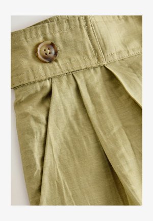 Nahaufnahme von beigen Stoffhosen mit einer plissierten Vorderseite und einem braunen Vierlochknopf am Bund.