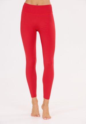 Røde leggings med høj talje i fuld længde, båret af en person, der står barfodet på en hvid baggrund.