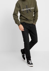 Person wearing olive green Calvin Klein Jeans sweatshirt, black slim-fit jeans, and black low-top sneakers with white laces and star logo.