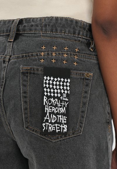 Black denim jeans featuring cross-stitching on the back pocket, with a black label displaying white text: "ROYALTY HEROISM AND THE STREETS."