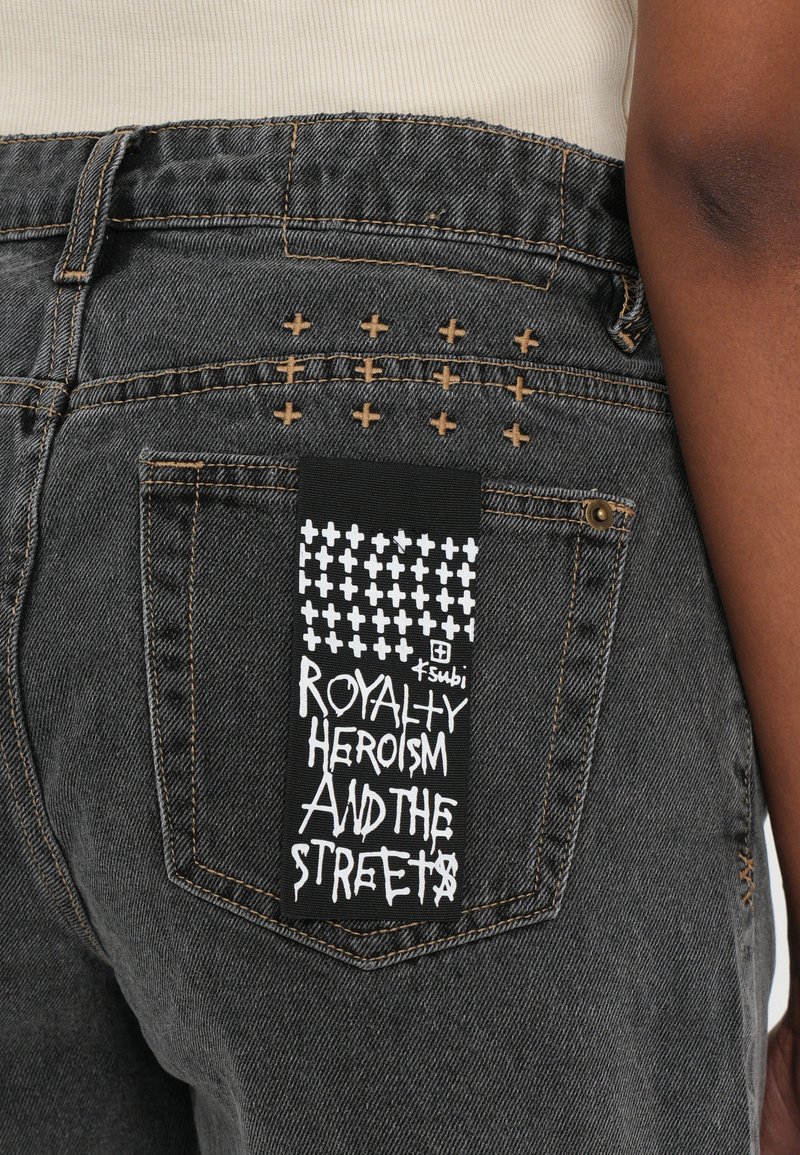 Jeans in denim nero con cuciture incrociate sulla tasca posteriore, dotati di un'etichetta nera con testo bianco che recita: "EROISMO REALTA E LE STRADE."