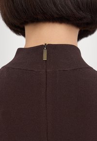 Brun stickad plagg med hög krage och en kort dragkedja på baksidan, med en liten metalltagg nära dragkedjehandtaget.