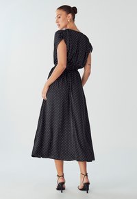 Schwarzes Midi-Kleid mit weißen Punkten, kurzen Ärmeln, geraffter Taille, glatter Textur und lockerer Passform; kombiniert mit schwarzen Riemchen-Heels.