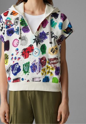 Haut à capuche blanc à manches courtes avec des dessins abstraits colorés et des griffonnages, porté avec un pantalon vert olive sur une personne sur fond gris.