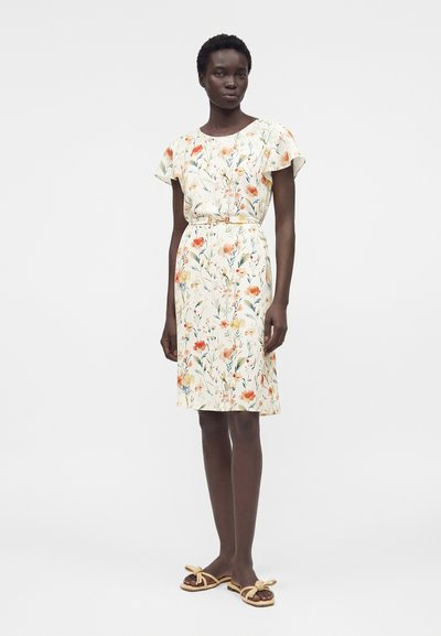 Γυναίκα που φοράει ένα φορέμα με κοντά μανίκια και φλοράλ σχέδιο μέχρι το γόνατο με ζώνη, συνδυασμένο με μπεζ σανδάλια με φιόγκο, στέκεται μπροστά σε απλό φόντο.