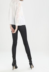 Blusa bianca con maniche in pizzo, abbinata a jeans skinny neri e stivali con tacco neri, che mettono in mostra una silhouette aderente e texture morbide.