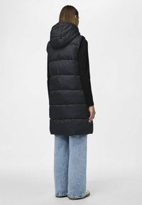 Gilet imbottito di un profondo blu navy con cappuccio, caratterizzato da un design trapuntato, cuciture laterali e una lunghezza maggiore, indossato sopra una maglietta nera a maniche lunghe.