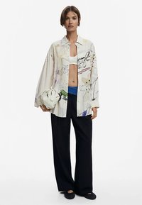 Camicia a maniche lunghe e larga con motivi floreali in tonalità morbide, indossata sopra un top corto bianco e abbinata a pantaloni neri ampi.