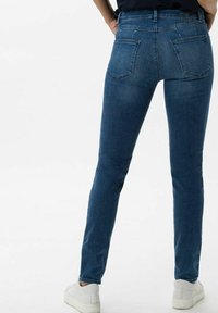 Vaqueros ajustados de mezclilla azul oscuro, con cintura media, bolsillos traseros y textura suave; combinados con zapatillas blancas.