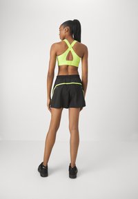 Sujetador deportivo en verde neón con tirantes entrecruzados y detalles recortados, combinado con pantalones cortos negros que tienen acentos en neón y una cintura elástica.