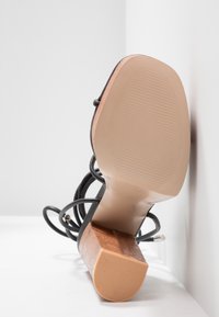 Sandale noire à talon unique avec lanières nouées autour de la cheville, semelle beige et talon bloc en bois, posée contre un coin blanc.