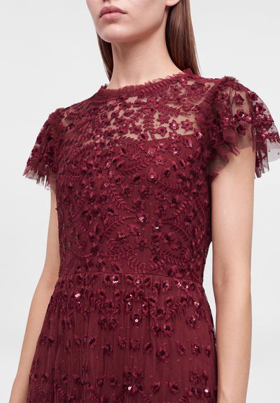 Femme portant une robe bordeaux avec broderie en dentelle florale, sequins et manches bouffantes courtes transparentes sur un fond uni clair.