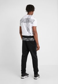 White t-shirt with black text pattern, paired with black sweatpants featuring a text-accented waistband, worn with black shoes.