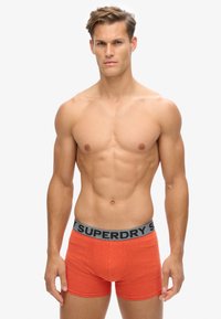 Boxer ajustados de algodón rojo con una cinturilla elástica gris que presenta el logo "SUPERDRY". Diseño ajustado y textura suave. El modelo está de pie, de frente.