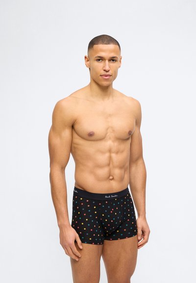 Αθλητικός άνδρας που φοράει μαύρα μποξεράκια με πολύχρωμες πουά, στέκεται μπροστά σε απλό γκρι ανοιχτό φόντο.