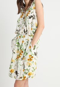 Robe sans manches à motifs floraux sur fond blanc avec des motifs jaunes et verts, taille élastique et poches latérales.