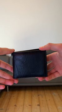 Zwarte leren portemonnee met een gestructureerde afwerking; bi-fold ontwerp, rechthoekige vorm, zichtbare stiksels, vastgehouden tussen twee handen tegen een eenvoudige achtergrond.