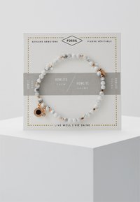 Valkoinen howlite-helmi rannekoru kultaisilla yksityiskohdilla, jossa on musta charm ja koristeellinen pakkaus. Rannekoru on pyöreä ja siinä on sileä pinta.