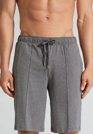 Persona sin camiseta usando pantalones cortos grises con cordón, de pie con los brazos relajados a los lados sobre un fondo liso.