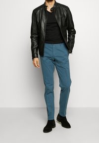 Zwarte leren jas met opstaande kraag, gecombineerd met lichtblauwe broek. Model draagt een zwart T-shirt en zwarte suède enkellaarsjes.