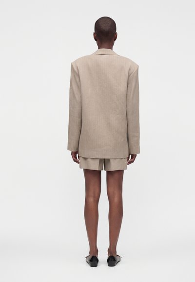 Άτομο με κοντά μαλλιά που φοράει μπεζ oversized σακάκι και ταιριαστό σορτς, όρθιο με την πλάτη σε απλό ανοιχτό φόντο.