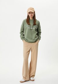 Sudadera verde con capucha y letras "PASADENA CALIFORNIA", pantalones de pierna ancha beige, zapatillas blancas y una gorra color tostado. Texturas de tela suaves.