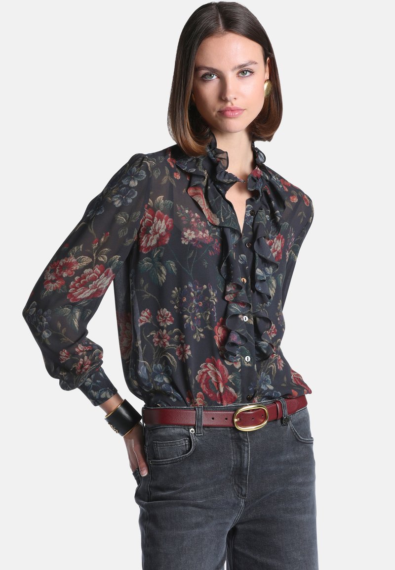 Femme aux cheveux bruns mi-longs portant un chemisier noir transparent à fleurs avec des volants, un jean foncé, une ceinture rouge et des boucles d’oreilles en or.