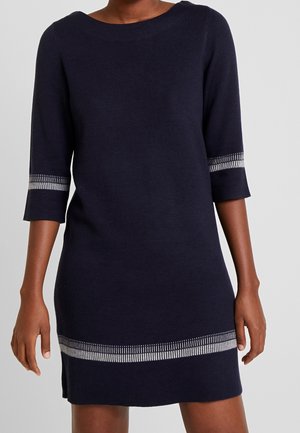 Modèle portant une robe bleu marine foncé à manches trois-quarts avec des détails en argent aux poignets, positionné devant un fond blanc uni.