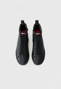 Une paire de baskets hautes en cuir noir avec des lacets noirs et un logo Camper rouge sur la languette, vue de dessus sur un fond blanc.