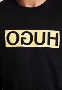 Černé tričko s žlutým obdélníkovým logem přes hruď, na kterém je v obrácených černých velkých písmenách napsáno "HUGO".